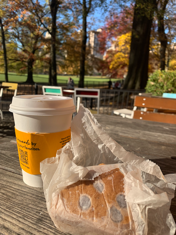 a coffee cup and a pastry sit on a table with trees and a field in the background.