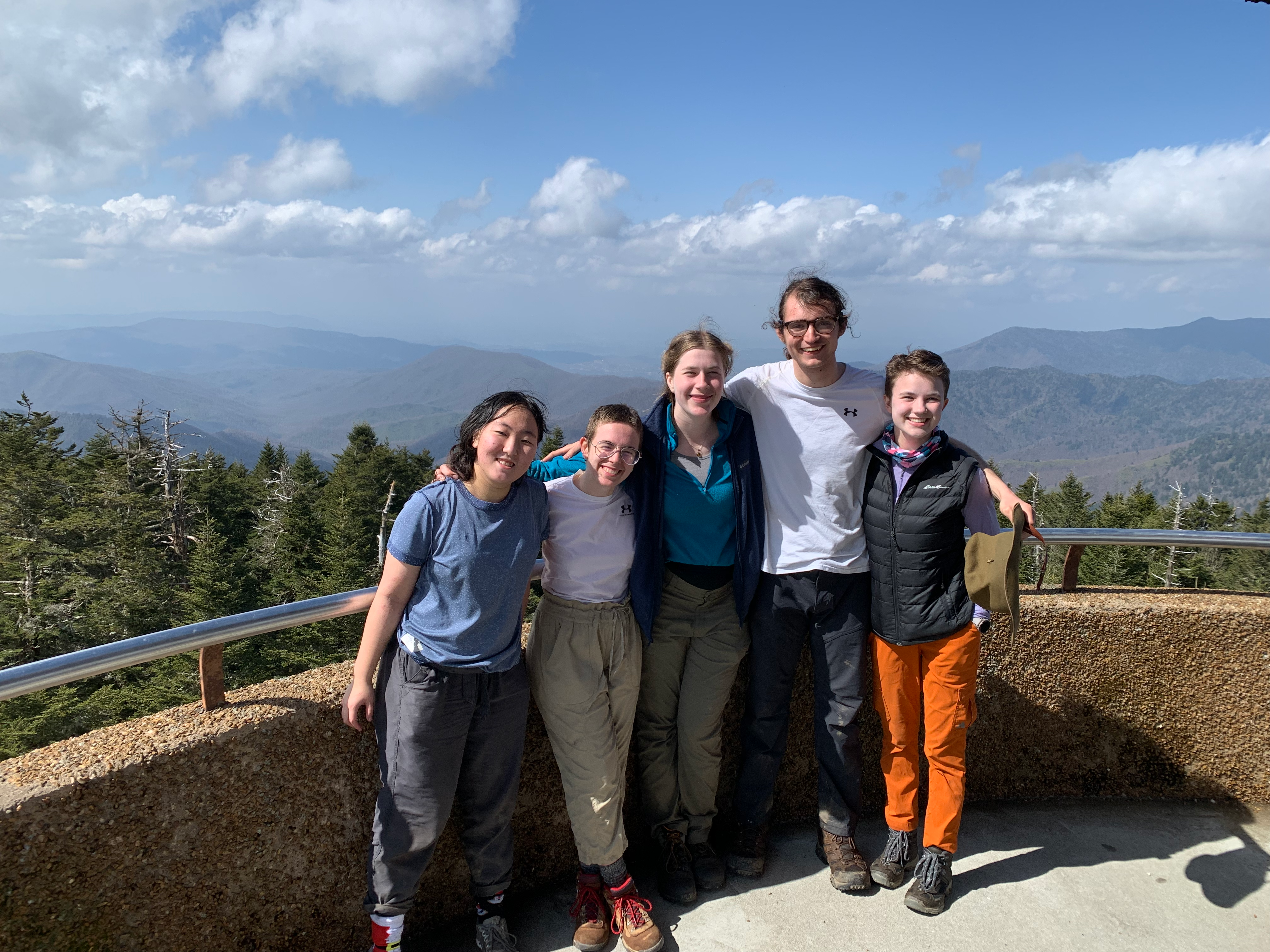 five smiling people stand at a ledge that overlooks lots of mountains