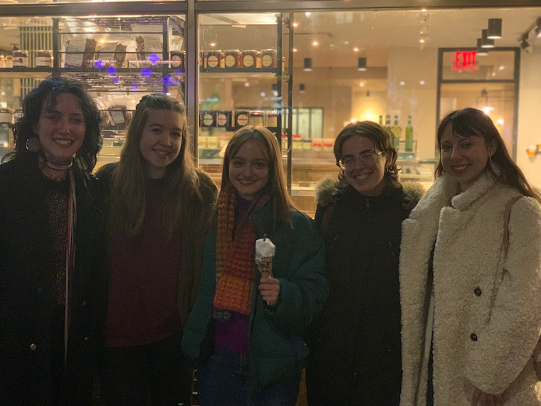 five people in coats smile at the camera outside of a store. one is holding an ice cream cone.
