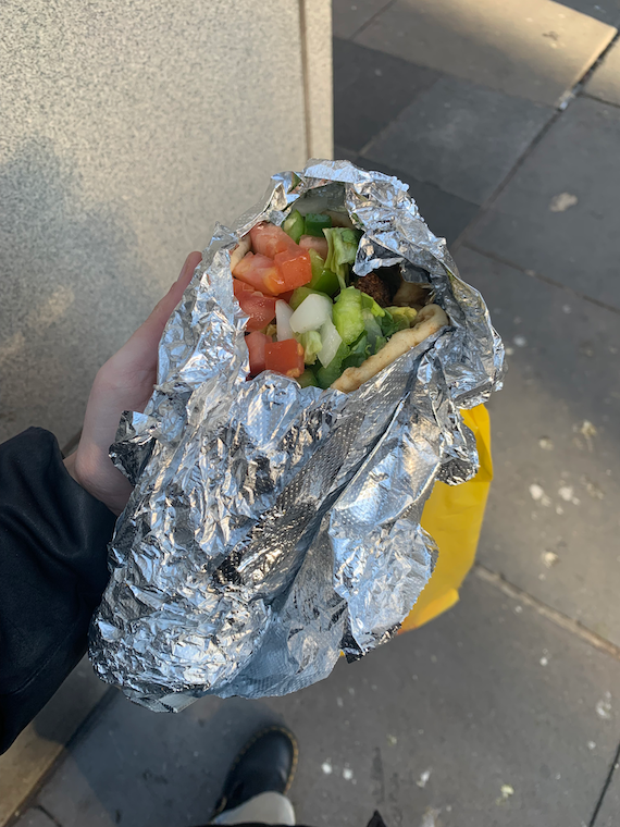 a foil-wrapped pita with tomatoes, lettuce, and other toppings is held in the street