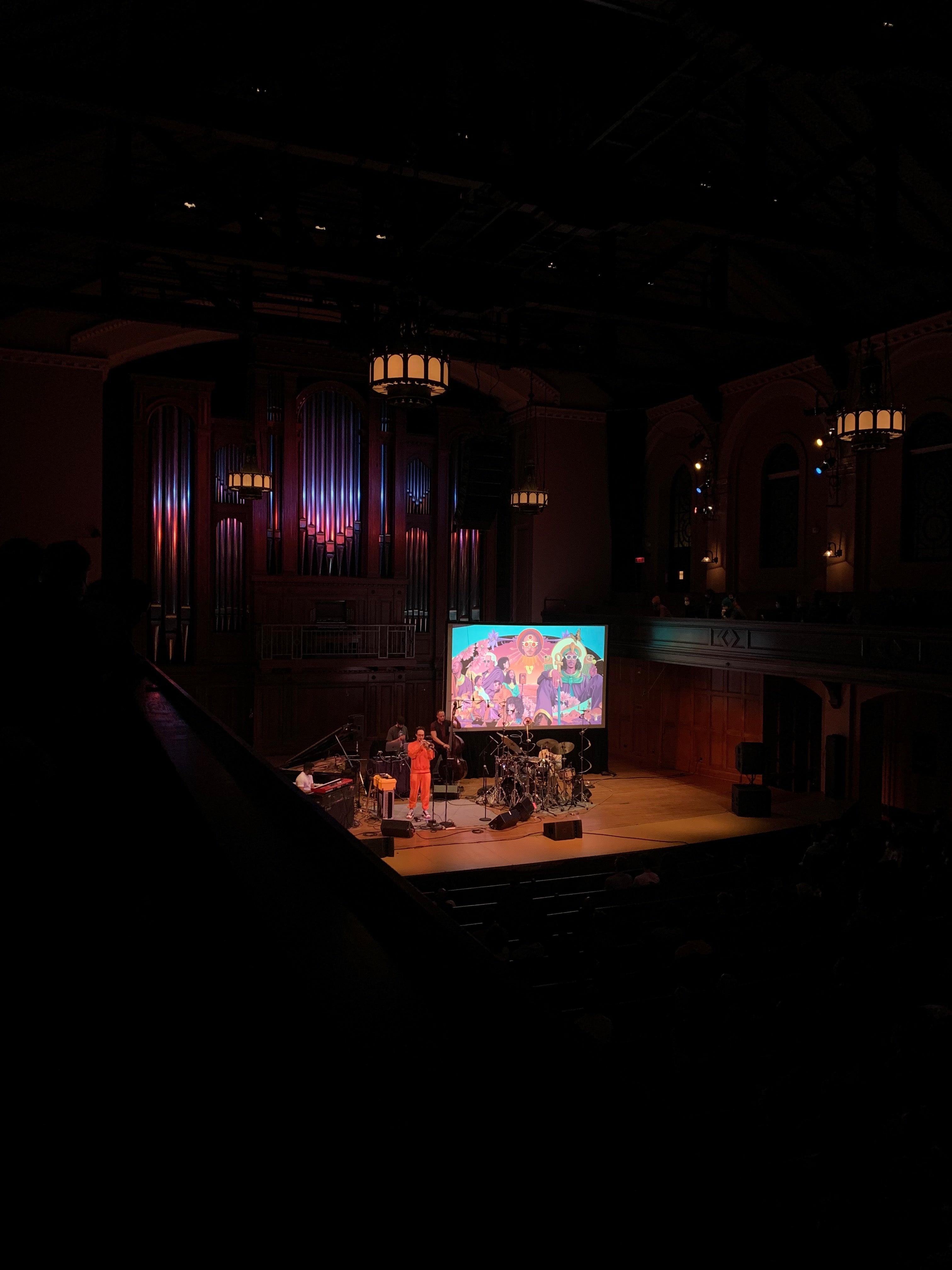 a trumpet player and band stand on the stage of Finney Chapel with a psychedelic backdrop behind them