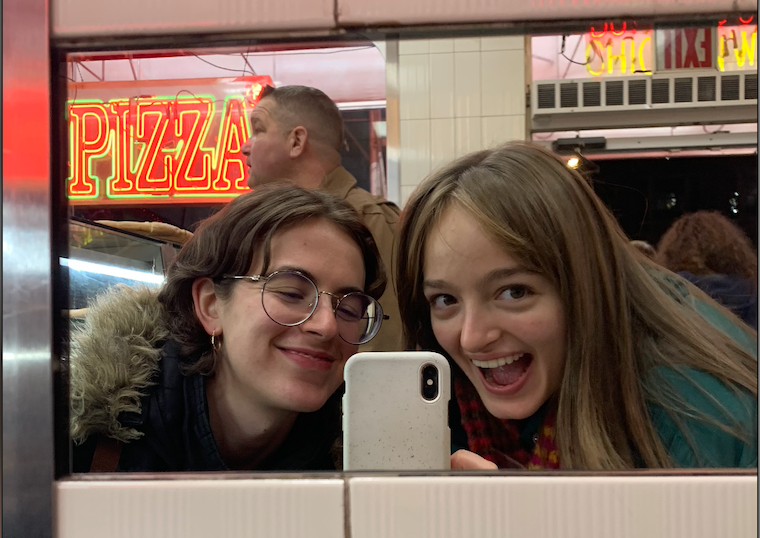Ilana and another person take a mirror selfie with a giant neon reading "PIZZA" in the background.