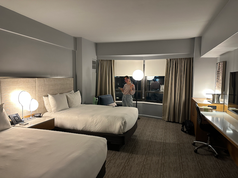 Ilana waves to the camera in a large and well-furnished modern hotel room.