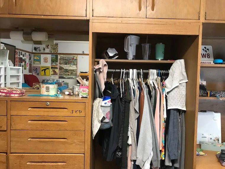 on the left is a wooden dresser with small boxes and hair accessories on top. Above it is a mirror in which you can see art on the opposite wall. On the right you can is a closet area filled with hanging clothes