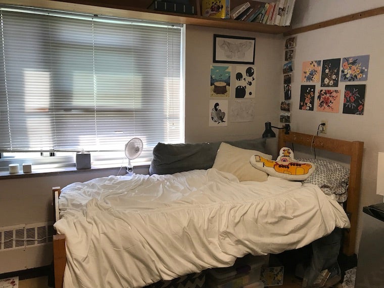 There is a wooden twin bed next to a window. The bed has a white comforter and several pillows. There is art hanging to the right of the window and above the head of the bed. There is a wooden shelf above the window filled with books.