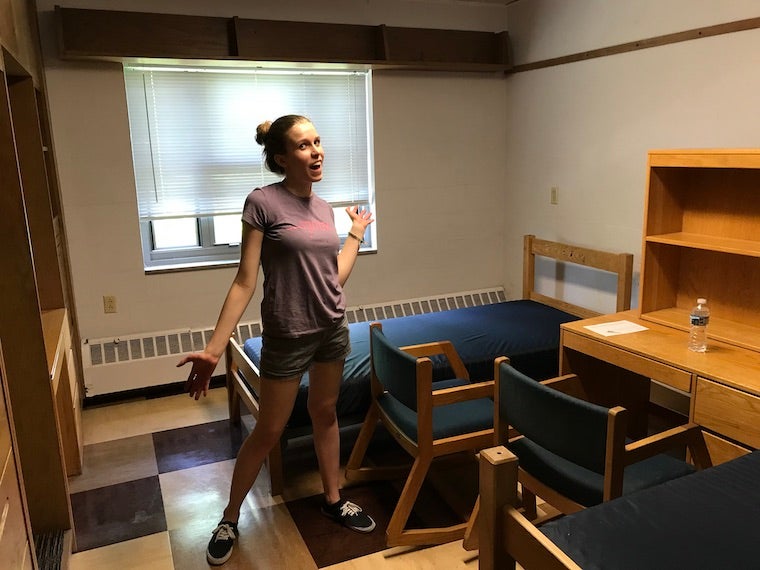 a photo of me standing in the middle of my dorm room. You can see a wooden twin bed with a blue mattress, a wooden desk, and two wood/blue fabric chairs