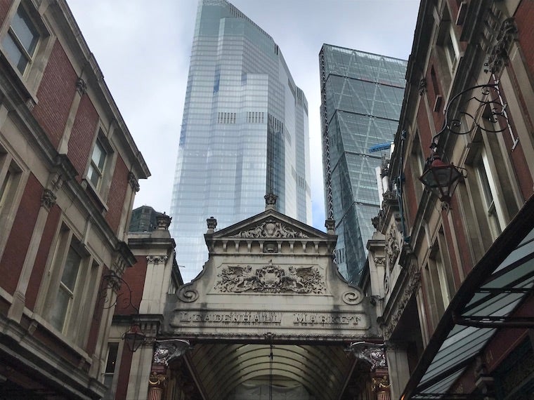 An old building with red brick and light colored stone. The words "Leadenhall Market" are carved into the stone. Behind, two big, shiny skyscrapers rise into the clouds