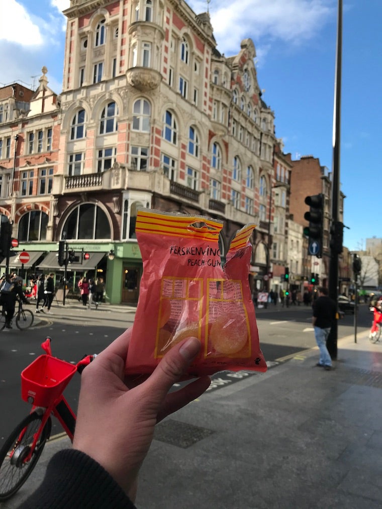 In the center is my hand holding a pink package of peach candy. In the background is a red bicycle, a sort of peach-colored building, and blue skies