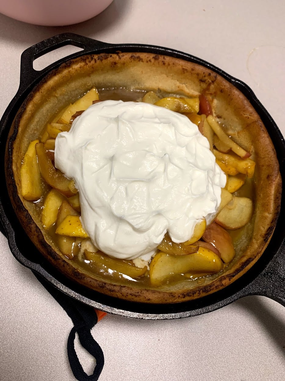 A crater dutch pancake sits in a cast iron skillet. Inside the dutch baby pancake there are cooked caramelized apples topped with whipped cream. 
