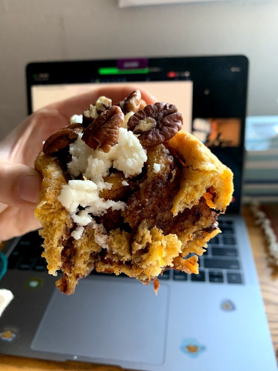 Half of an orange colored cinnamon roll with white icing and pecans is held in front of a computer screen on which a zoom class is occurring.