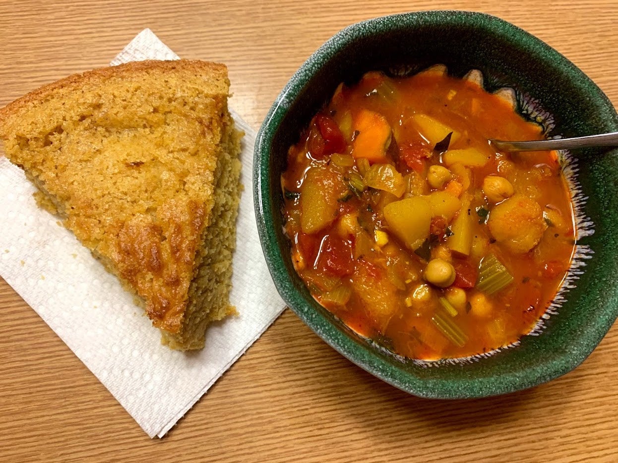 On the left, a slice of yellow cornbread sits atop a paper towel. On the right, there is red soup with many vegetables and chickpeas in a ceramic bowl with a spoon. 