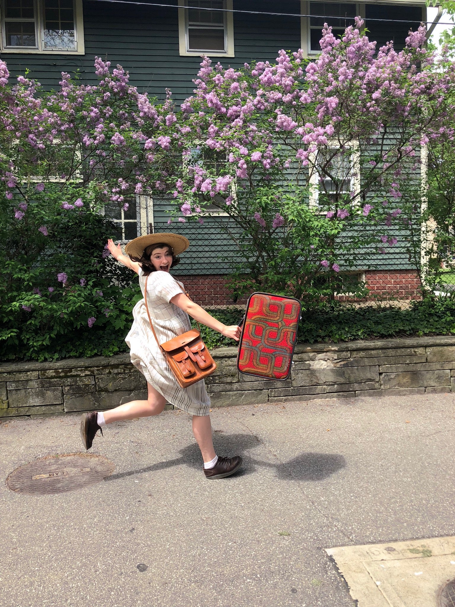 A girl wearing a hat and carrying a suitcase jumps in front of a bush with purple flowers 