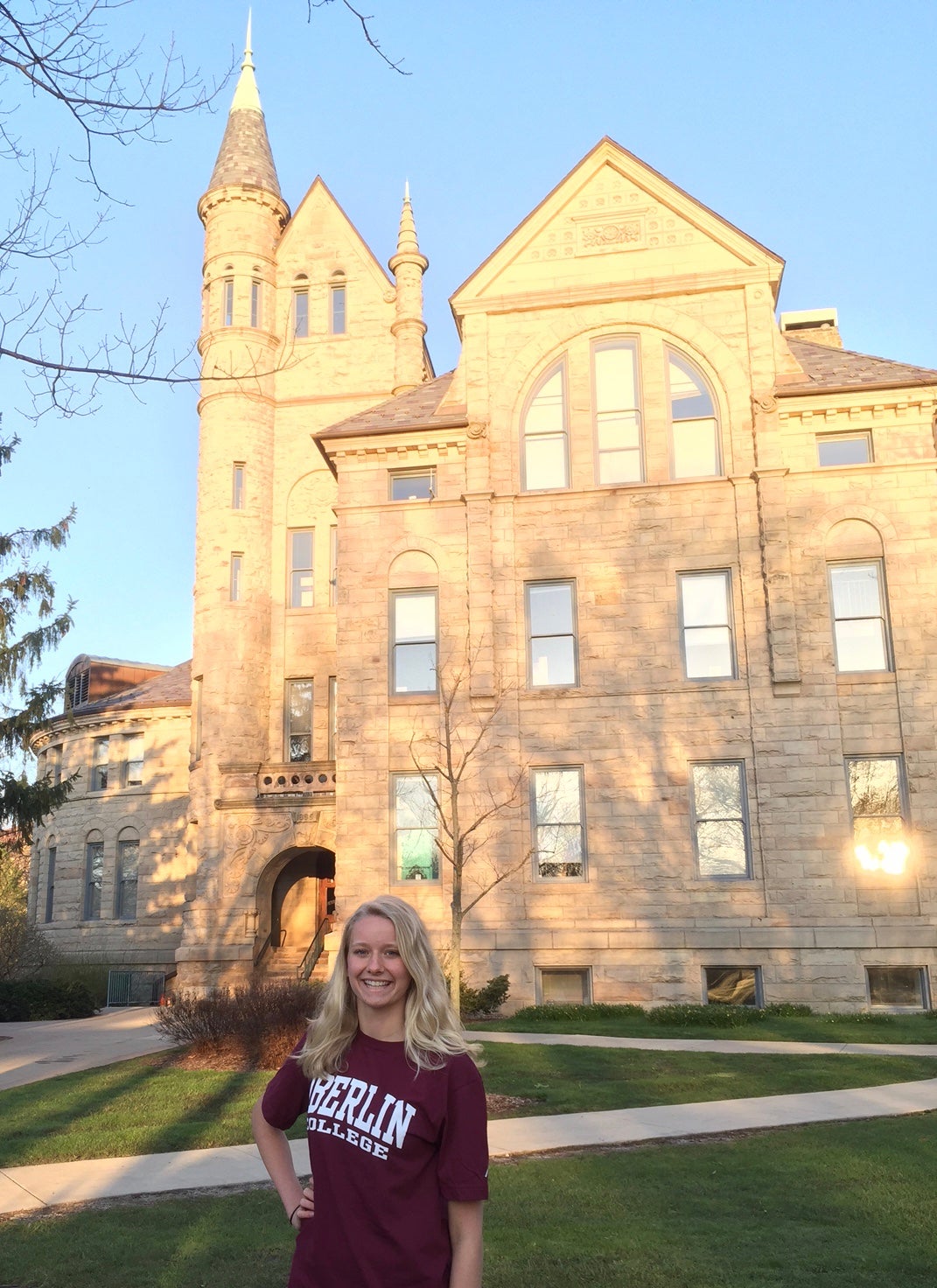Sarah stands in front of Peters building on Oberlin campus, wearing an Oberlin shirt