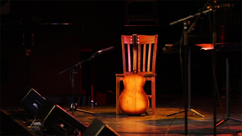 An acoustic guitar rests against a chair on a lighted stage with microphone stands and speakers.