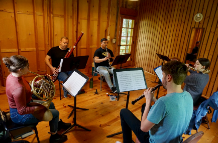 A woodwind quartet with Bebe playing horn rehearses in a cabin