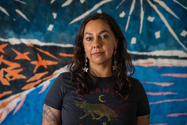 A portrait of Tongva artist Mercedes Dorame wearing a dark graphic T-shirt and star-shaped earrings. Dorame looks directly at the camera with a calm, thoughtful expression. The colorful backdrop, with its bold blues, oranges, and white radiating forms, echoes themes of land, water, and sky that are central to the artist's practice.
