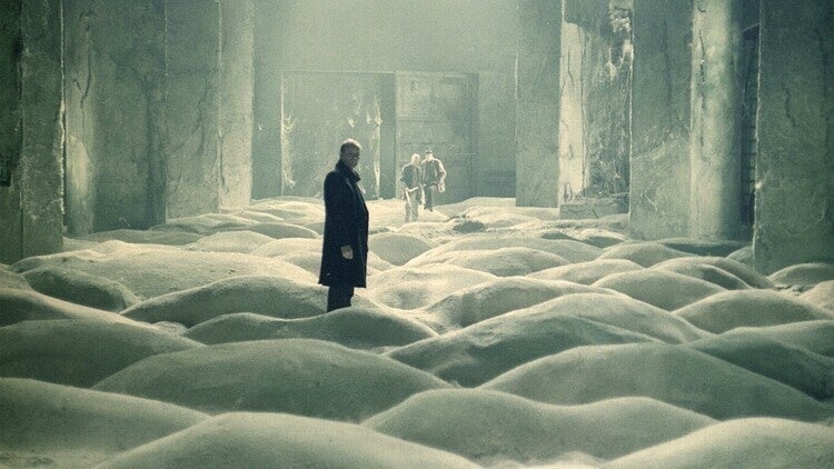 A green/grey underground room filled with mounds of sand. One man stands in the foreground, two in the back. 