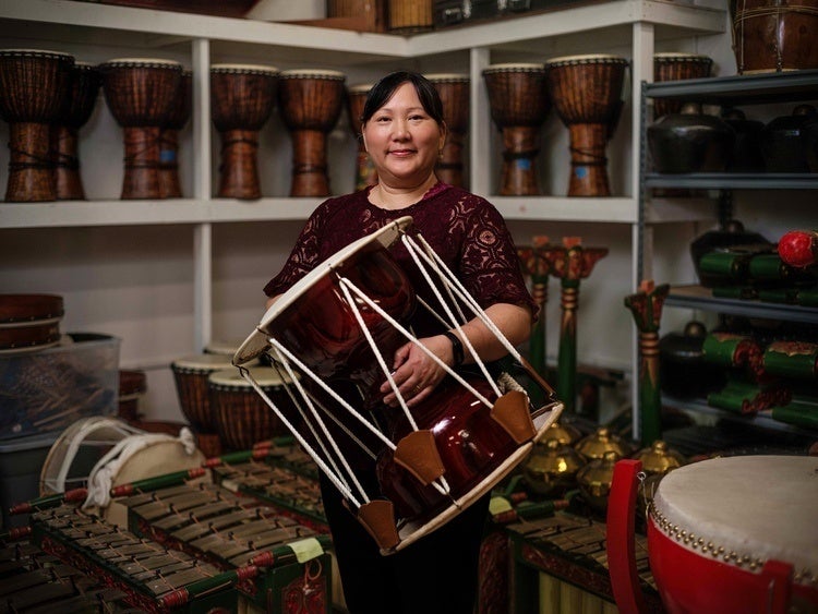 Dr. Donna Kwon stands in the center of a room holding a Korean drum, surrounded by shelves with drums