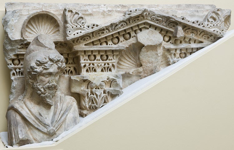 Aged carving of a figure wearing a hat against an architectural background.