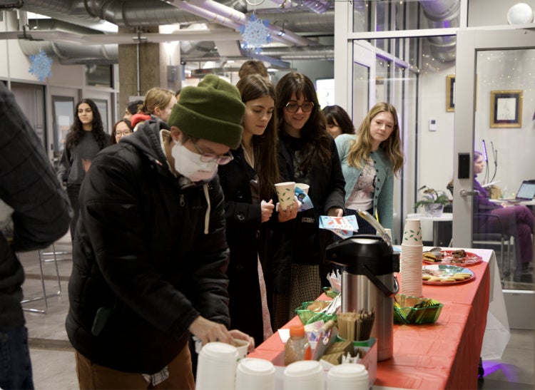 Students line up at a table pouring hot chocolate from a dispenser at an indoor social event.