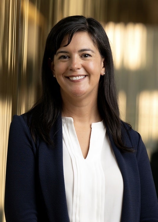 Woman with long dark hair, dressed in a navy blazer and white blouse, smiling at the camera.