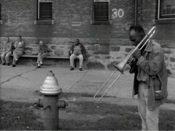 Inmate-patients sit outside on the sidewalk; one plays the trombone in the foreground