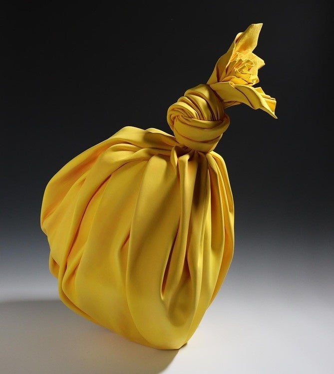 This photo is an image of a ceramic sculpture. The sculpture is a bright yellow bag that appears like cloth tied in a knot.