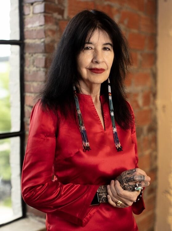 A person with dark hair in a red blouse and long beaded earrings stands in front of a brick wall.