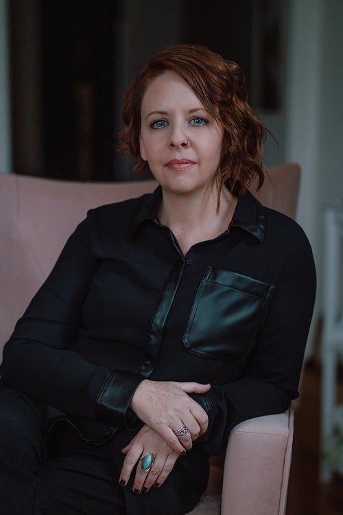 A person in all black leans on the arm of a muted pink arm chair and looks forward into the camera.