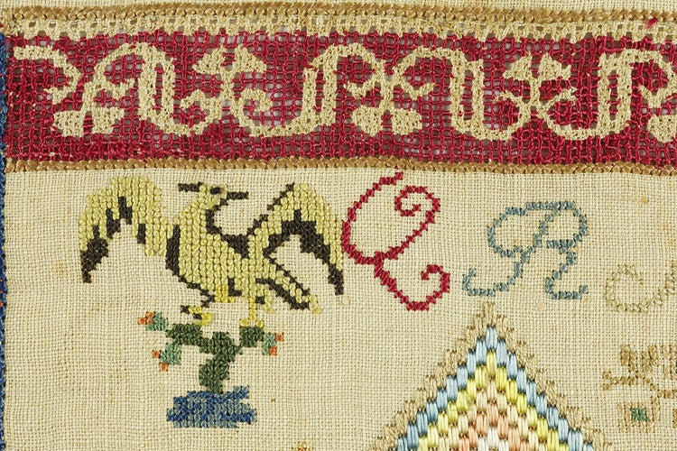 A zoomed in section of an embroidered piece featuring a red decorative strip of red with light yellow detailing at the top, a bird landing on a cactus, a red Q, and a blue R. 