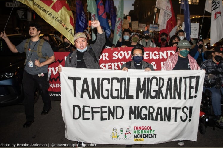 Tanggol Migrante Movement activists holding a sign that reads: "TANGGOL MIGRANTE! DEFEND MIGRANTS!"