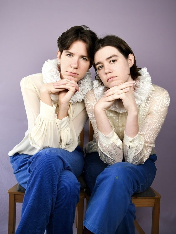 Two brown-haired people pose upright with hands clasped beneath their heads. They wear old fashioned ruffle collars and look at the camera.