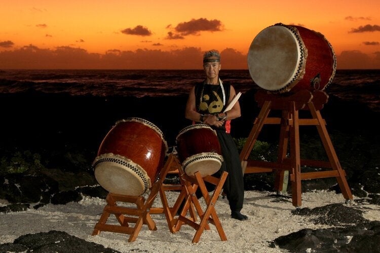 Kenny Endo stands on a beach at sunset, holding his bachi (taiko drum sticks). In front of him are two smaller taiko drums, on the left is a large taiko drum. 