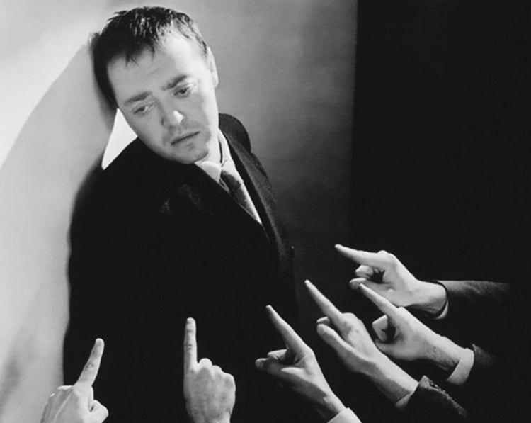 Black and white photo of several hands pointing at an upset man on the wall. 