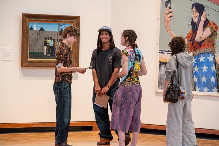 Four students stand in an art gallery, three stand together in discussion as one looks at a painting off to the far right.
