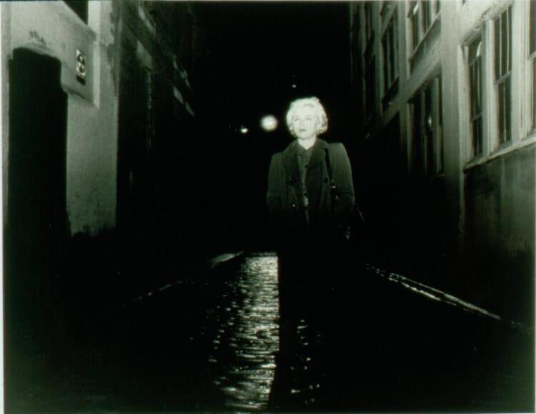 The photo is a black and white image of a woman with short hair standing in an alley.
