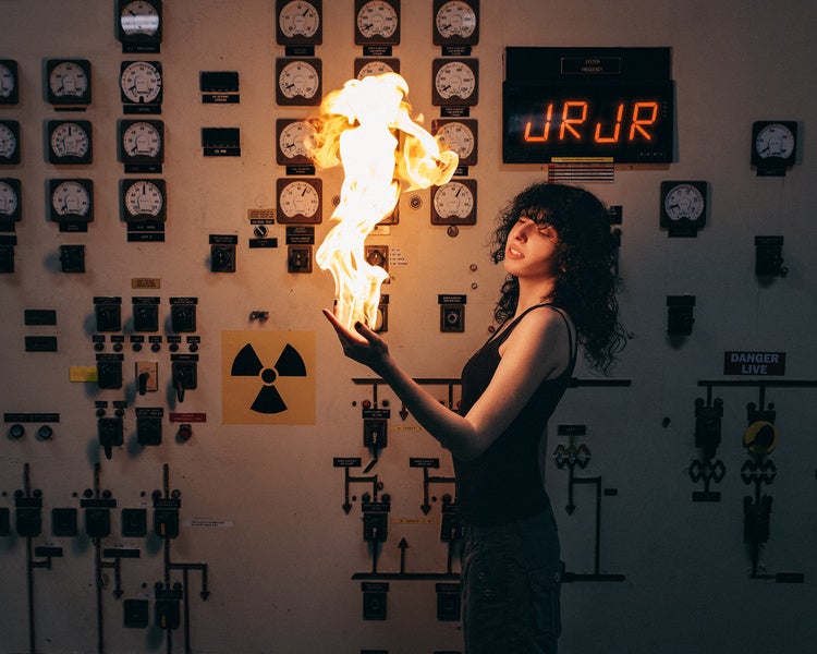 Person standing in front of a control panel with gauges and switches, holding a large flame in one hand near a radiation warning sign.
