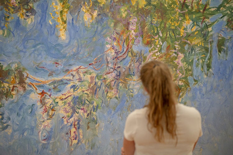 Photo from behind of student looking at blue painting with colorful, gestural brush strokes, concentrated in the upper right corner of the piece.