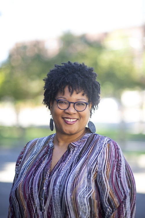 Smiling woman with short curly hair, glasses, earrings, and a striped blouse, standing outdoors.