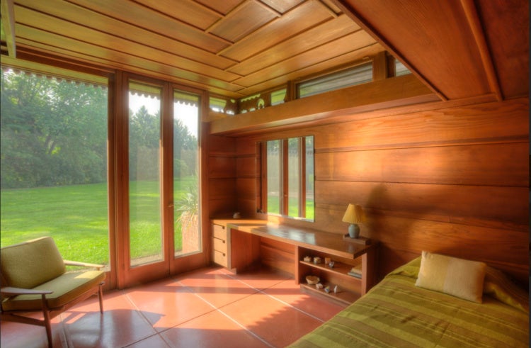 A warm, wood-paneled room with large windows overlooking a green lawn, featuring a built-in desk, armchair, and bed in golden sunlight.  