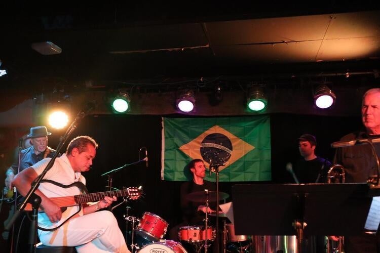 Brazilian singer Moises Borges on-stage at the 'Sco playing guitar in a chair with a microphone in front of him. Drummers are featured in the back. Brazilian flag hangs from wall. 