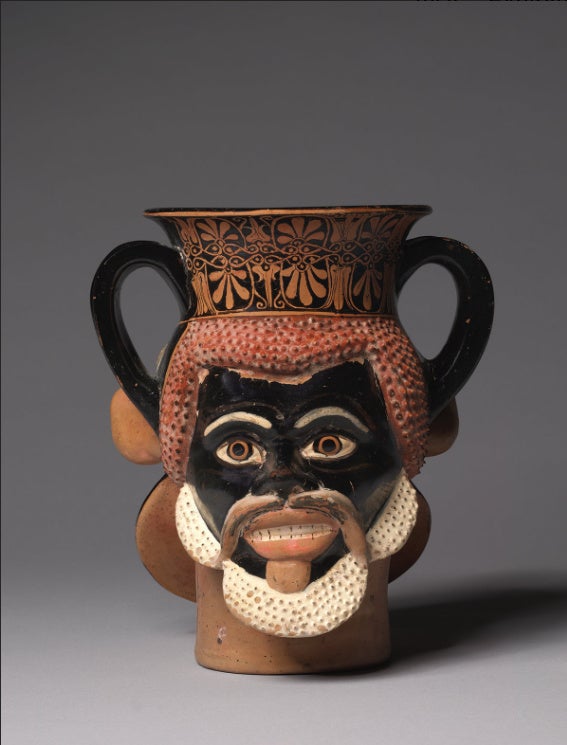 An image of an ancient Greek drinking cup with handles on both sides. The cup resembles a bearded face and has ornamental trim around the opening.