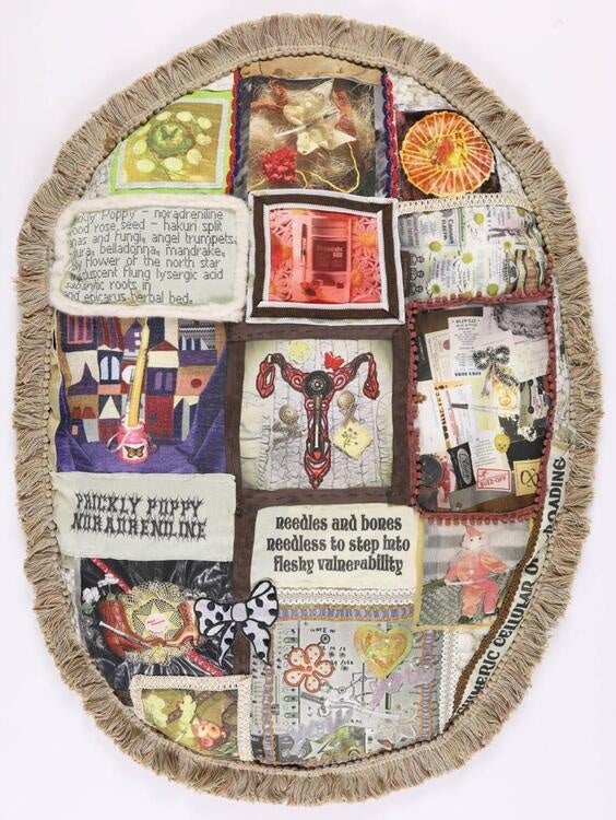 This photo is an image of a ovular pillow with brown tassels around the edges. The body of the pillow has images of a uterus, words, and a bow. 