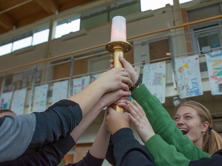 Hands of several people raised together holding a lit ceremonial torch indoors, with a smiling person in the background.