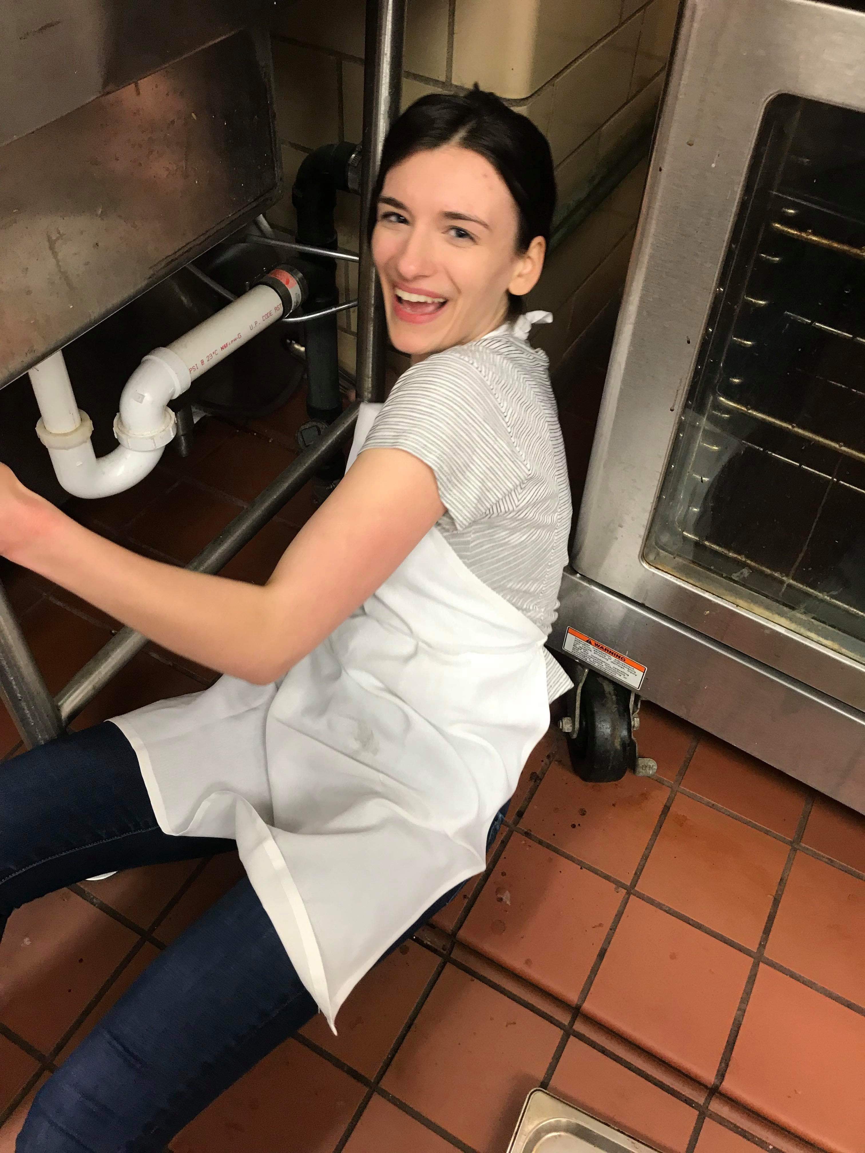 Megan sits on the red tiled floor of Fairchild Co-op's kitchen, wearing an apron and her hair in a ponytail. She is between an oven and a sink; her body is oriented toward the sink, a pair of tongs barely visible in her right hand, but she turns back to look at the camera. This is the happiest she has ever looked.