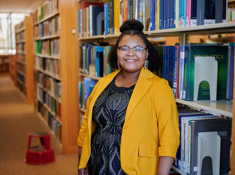 Email student standing in front of library book stacks wearing glasses and yellow blazer