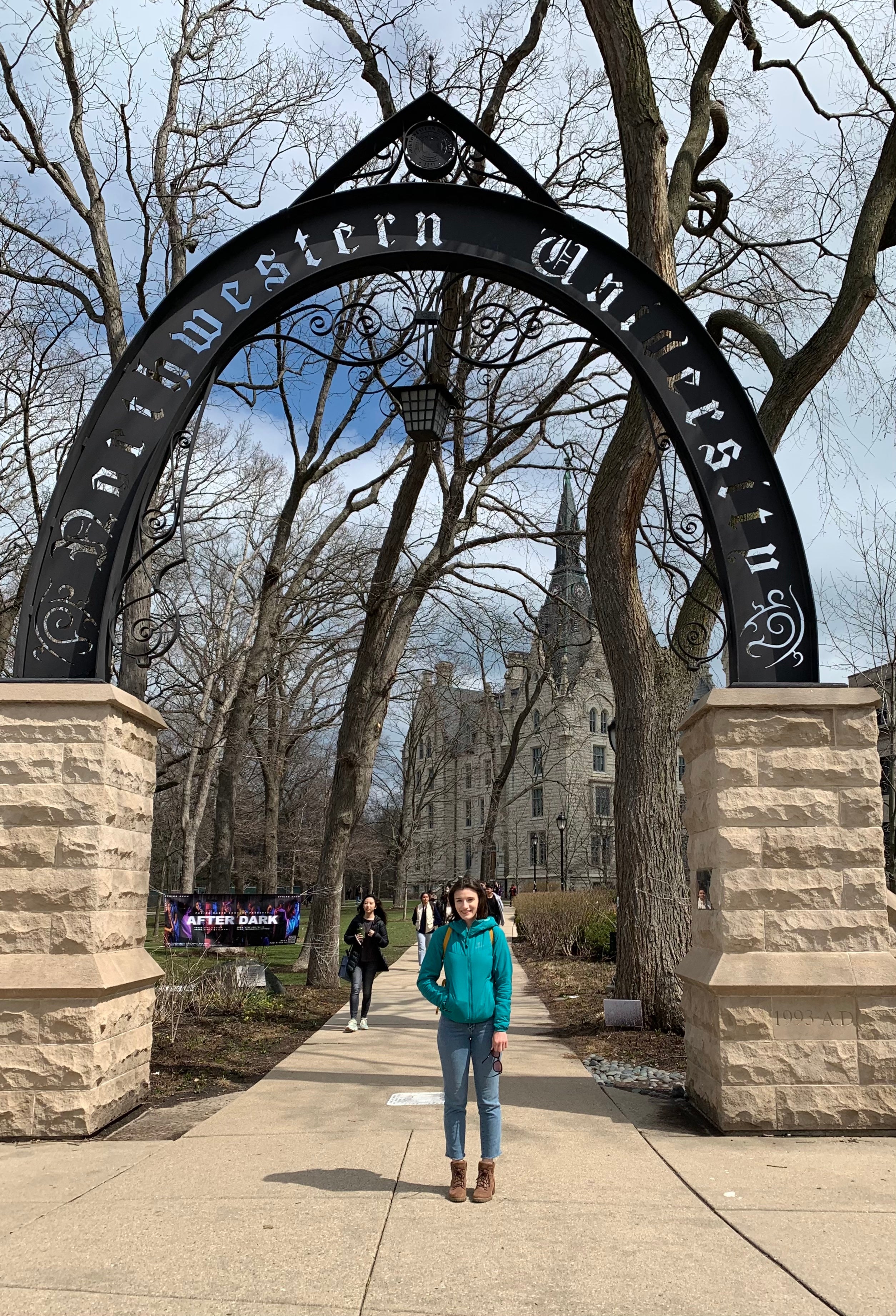 Megan stands in front of a stone and iron arch that reads "Northwestern University." It is a sunny day. She wears a light jacket and has one hand in her pocket while the other is at her side holding a pair of sunglasses. She is smiling.