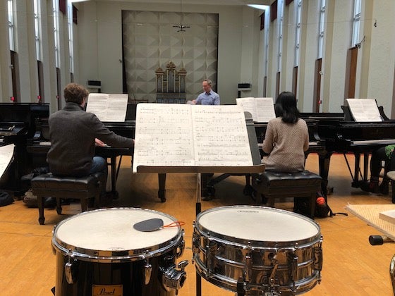 Two pianos, two drums, and a conductor in a concert hall.