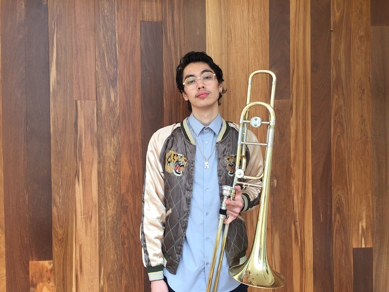 Photo of Neko Cortez posing against a wood paneled backdrop, holding a trombone