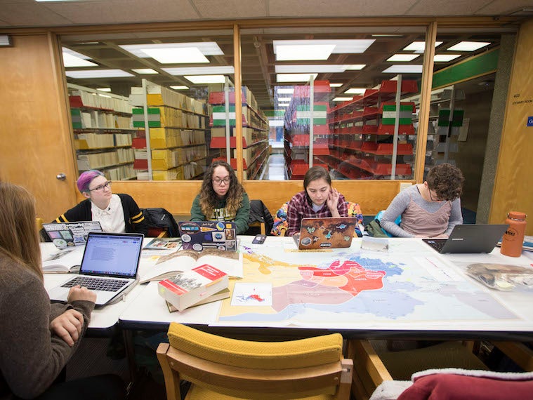Students sit at a table filled with maps and books.